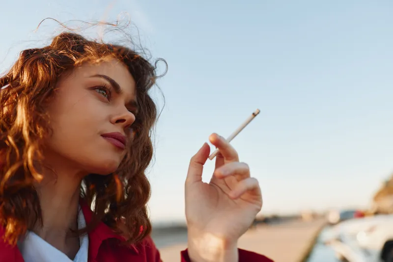 A woman with curly hair in a red jacket smokes a cigarette outdoors, gazing to the side in warm late afternoon light.