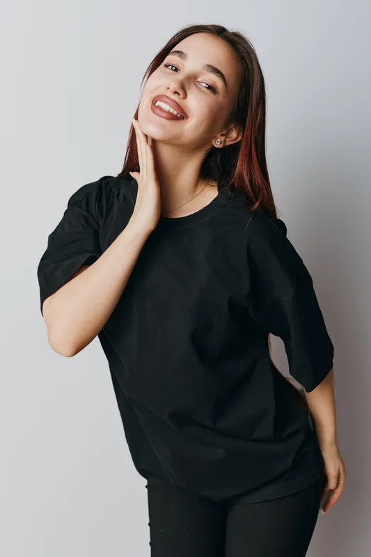 Young woman smiling and posing with hand on neck wearing casual black t shirt and pants against gray background in studio for lifestyle portrait and fashion concept.