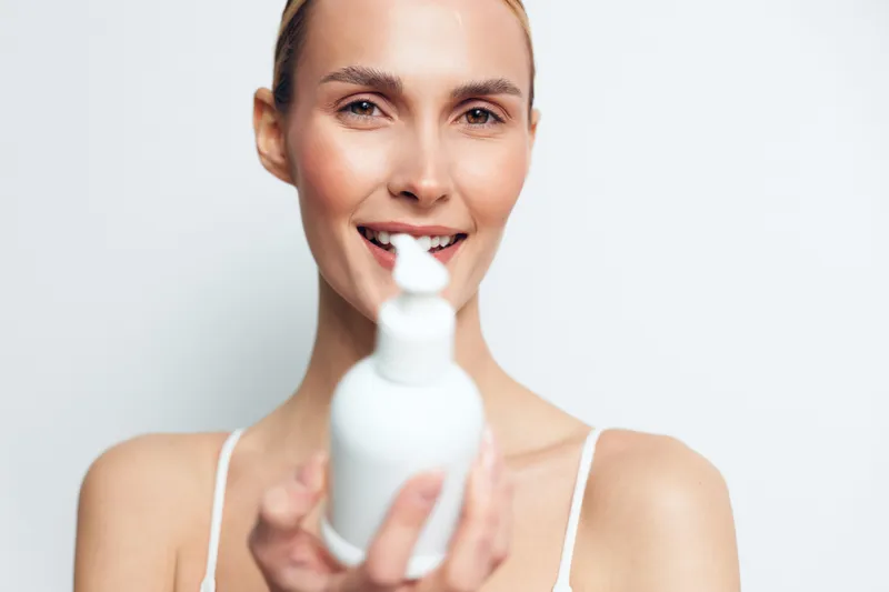 Young woman applies lotion from a pump bottle in a bright studio, smiling at the camera against a clean white background, capturing a fresh skincare moment of self care