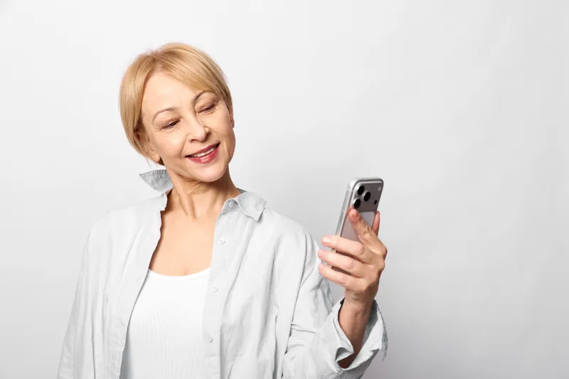 Smiling elderly woman holding smartphone for a selfie, cheerful communication technology portrait, casual studio shot isolated on light gray background clean