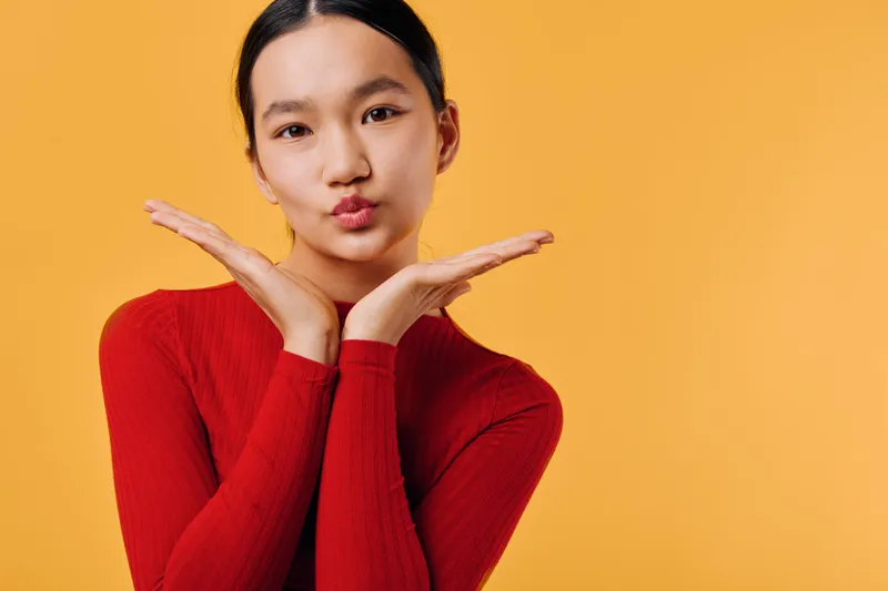 Young woman with dark hair wearing red sweater making playful face with hands under chin against orange background. Female portrait with casual expression and closeup on warm color wall.