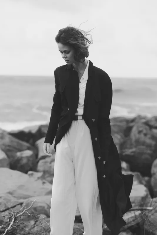 Windy coastal fashion scene with a stylish woman in a long dark coat and white wide pants, standing on rocky shoreline, moody monochrome tones and dramatic breeze.