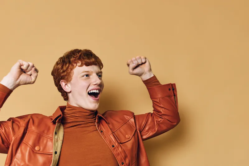Young man with red hair wearing brown jacket and turtleneck celebrating with raised fists against beige background. Enthusiastic, happy and excited adult male portrait with expression of joy.