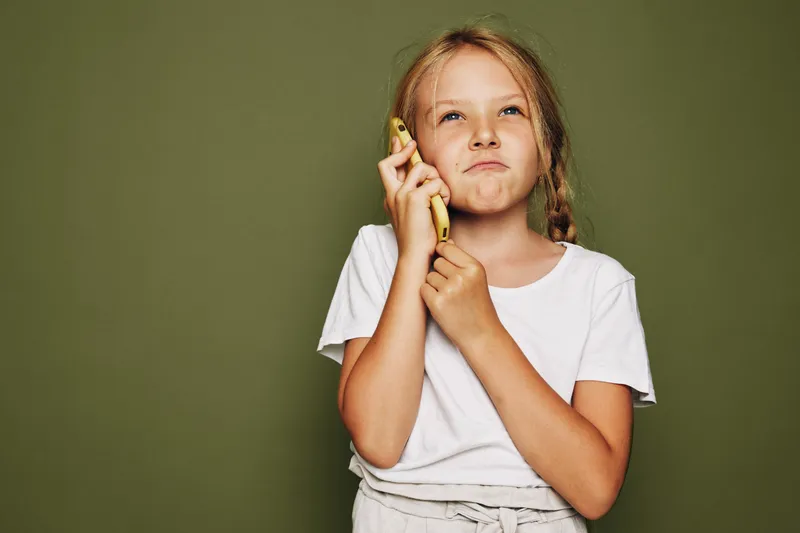 Portrait of a thoughtful girl holding a banana near her ear as if talking to a phone on olive background.