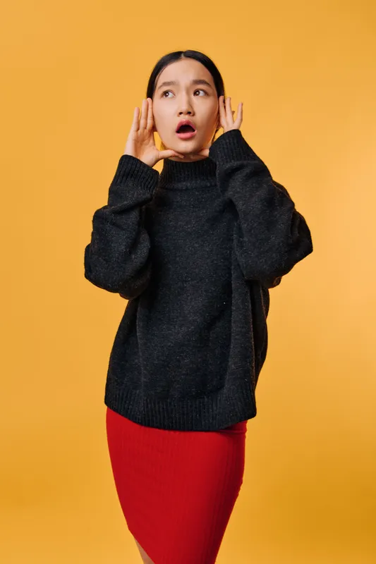 Woman with hands near ears and surprised face expression standing on yellow background wearing black sweater and red skirt. Young female showing shock or disbelief in casual outfit.