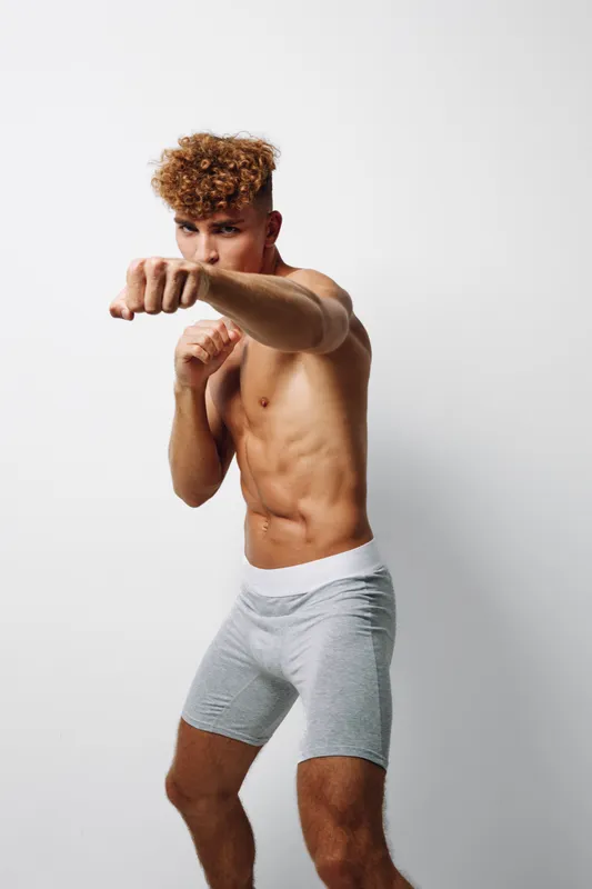 Athletic young man with curly hair in gray shorts practicing boxing stance and punches for fitness training and workout on white background in studio. Sport, exercise and strength.
