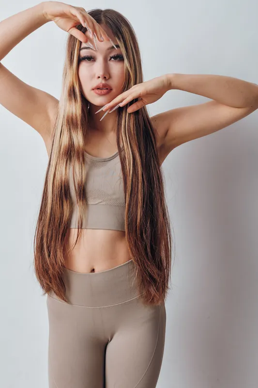 Young woman with long straight hair posing in beige sportswear against white background. Natural makeup, casual fitness outfit, confident and graceful with hands framing face and hair.