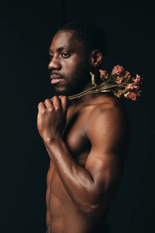 African man with dark skin holding dried flowers on shoulder in creative conceptual visual style. Portrait of shirtless male model with artistic expression on black background.