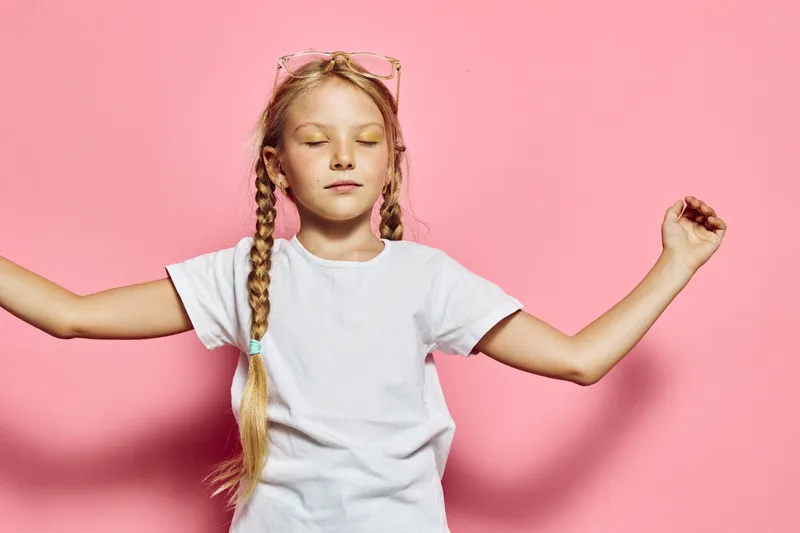 Blond girl with braids stands with closed eyes and raised arms against pink background. She wears a white t-shirt and light pink sunglasses on her head. Concept of calm, balance, and childhood.