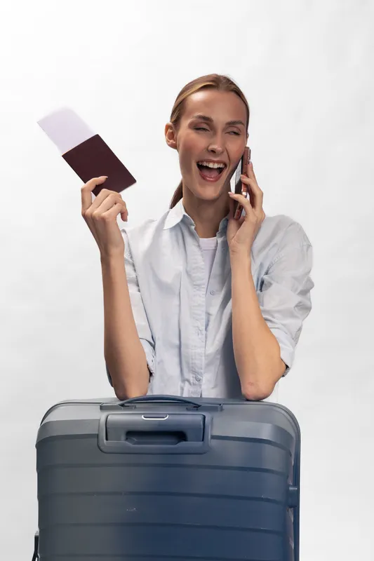 Happy young woman holding passport and tickets talks on phone, smiling excitedly behind large suitcase. Concept for travel, vacation, booking and holiday planning.
