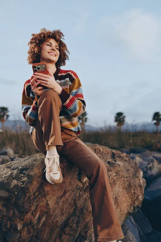 Woman lifestyle outdoor, curly hair woman sitting on a rock holding a smartphone, casual sweater and jeans, relaxed smile, soft evening light, nature background.