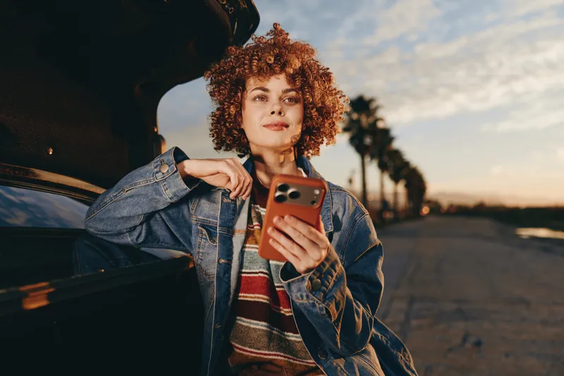 Curly haired woman lifestyle portrait in denim jacket and scarf holds a smartphone while leaning on a parked car at sunset, outdoors, warm light, calm mood, casual travel vibe