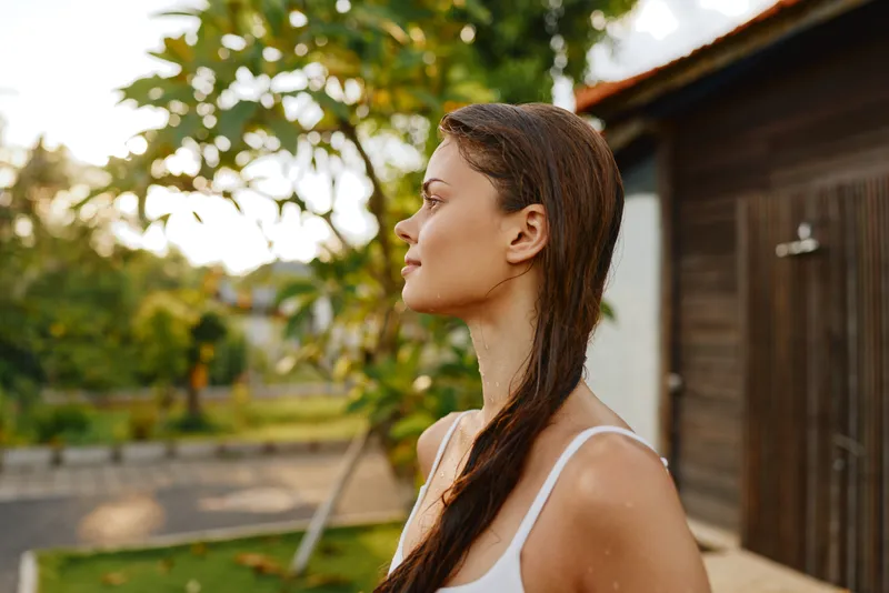 A woman in a white tank top stands in an outdoor scene, profile visible as sunlight touches her skin, surrounded by nature, trees and a wooden shed, conveying calm, casual lifestyle, serenity, and