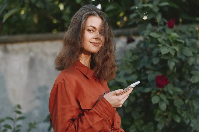 Cheerful woman outdoors, casual in an orange shirt, looking at her smartphone with a smile, sunlit garden backdrop, relaxed lifestyle moment