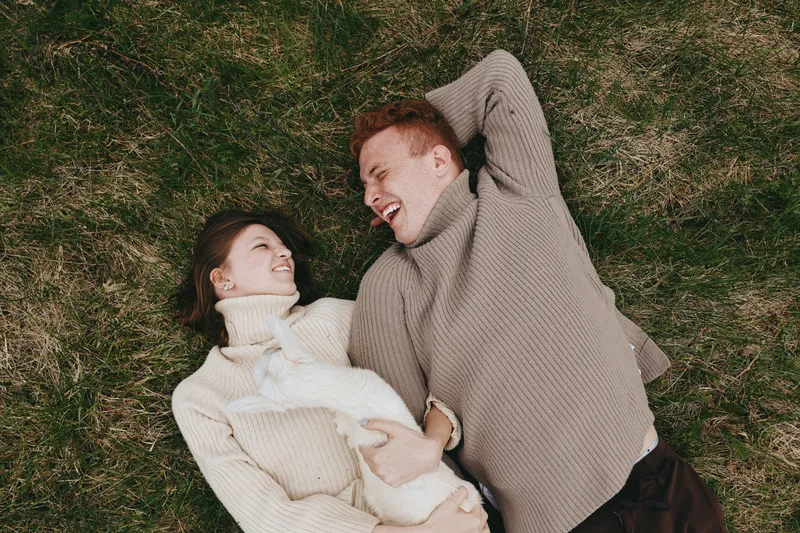 Young couple lying on grass wearing cozy sweaters while holding a white baby goat and sharing a joyful moment outdoors in a natural lifestyle setting.