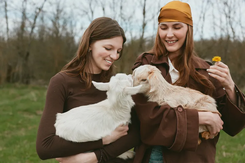 Two smiling women cradle a goat and a kid in a sunny field, capturing a warm farm lifestyle with gentle animal interaction, friendship, and care concepts for family life and wellbeing.