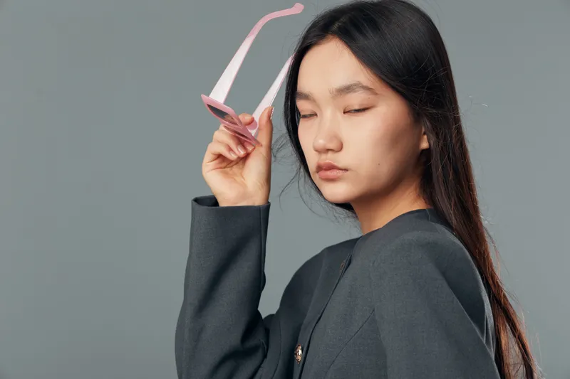 Young woman with long dark hair holding pink sunglasses, wearing dark jacket and eyes closed, posing on gray background in studio for fashion portrait or beauty concept.