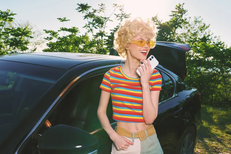 A stylish woman with curly blond hair in a striped crop top leans by a dark car, smiling at a phone. A bright retro mood and sunny lifestyle shine through a casual, colorful portrait.