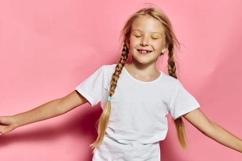 Smiling young girl with long braided hair wearing a white shirt stands with closed eyes and arms outstretched on pink background.