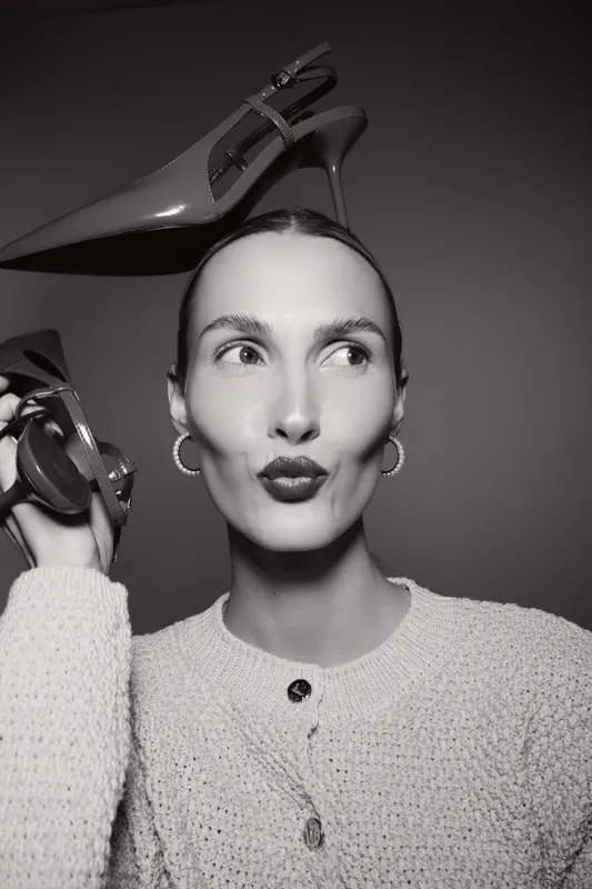 Portrait of a fair skinned caucasian woman with slicked hair holding high heel shoes near her head. Studio fashion concept with confident expression.