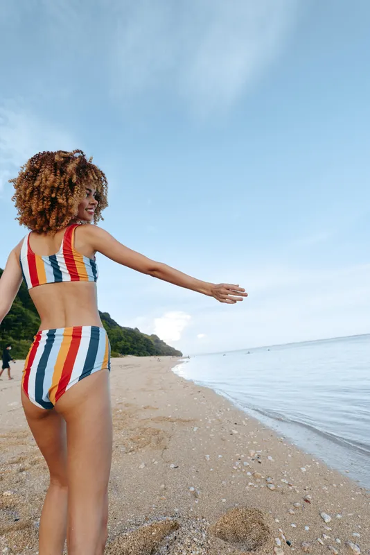 A young woman in a striped swimsuit stands on a sandy beach with an outstretched arm toward the sea, conveying a carefree summer moment and a sense of freedom.