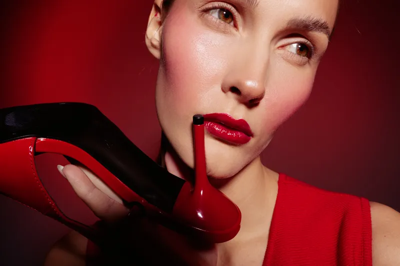 Close-up of a woman with red lipstick holding a red high heel shoe near her face. The image features bold makeup and fashion elements on a dark red background.