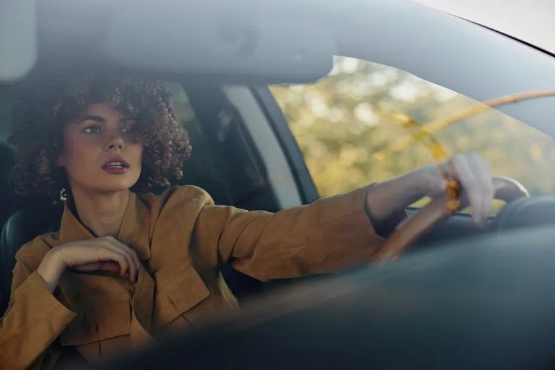 Woman driving a car with a brown jacket, looking to the side. Close-up on the steering wheel, warm light, retro film mood, everyday travel, candid and relaxed moment.