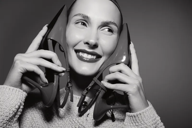 Happy Caucasian woman with fair skin smiles holding red high heel shoes near her face. Black and white studio portrait with cozy sweater. Concept for fashion and lifestyle.