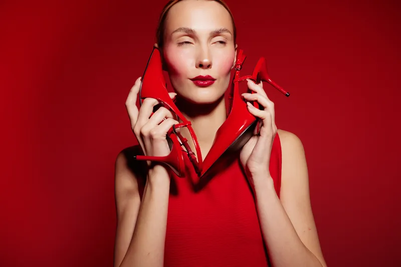 Woman in red dress holds red high heels near face with closed eyes on red background. Concept of fashion, style, elegance, and bold color contrast in studio lighting.