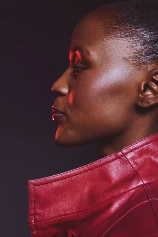 Profile of an African woman with dark skin wearing a red leather jacket. Studio portrait with dramatic lighting on a dark background. Concept of fashion, confidence, and elegance.