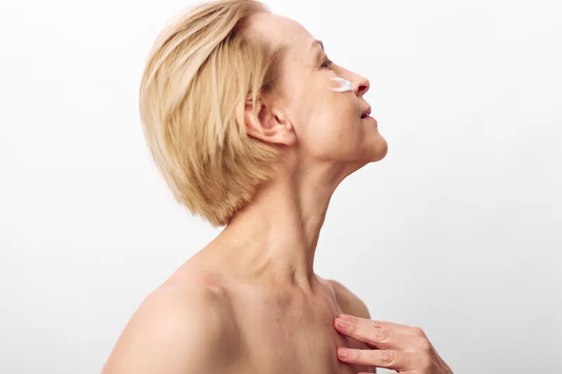 Profile of mature woman with short blonde hair applying cream under eye. Skincare concept focused on anti aging, hydration, and natural beauty on plain white background.