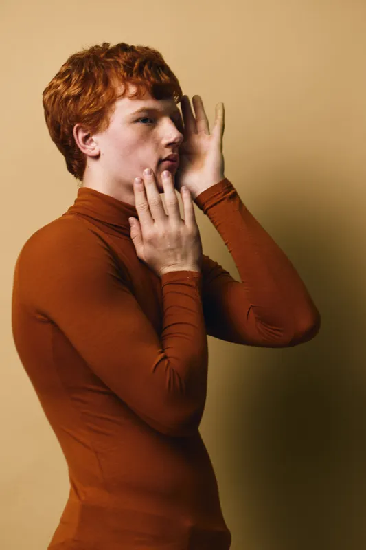 Young man with red hair in brown turtleneck posing with hands near face against beige background. Side profile portrait with soft lighting and neutral expression for fashion or art.