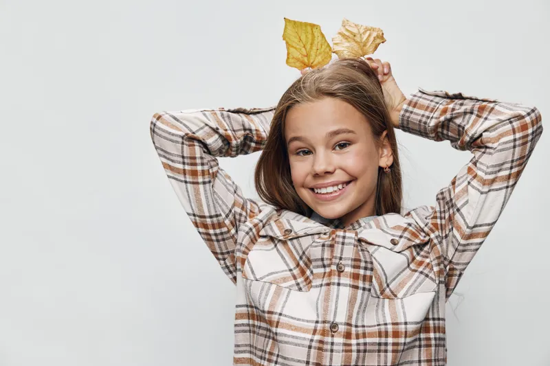 Smiling girl with brown hair wearing plaid shirt holding autumn leaves above head for fall season, joy and childhood concept isolated on light background.
