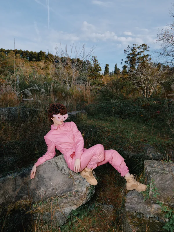 Woman in a pink suit lounging on a rock in an outdoor nature setting, autumn trees behind, relaxed pose, clear sky, fashion inspired portrait for lifestyle, travel and adventure themes