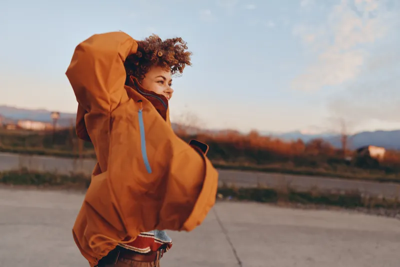 Woman with curly hair wearing hooded jacket outdoors in light rain, autumn lifestyle, walking on road near landscape at sunset, warm casual travel mood, soft wind and clouds today