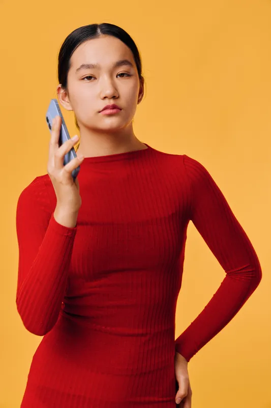 Young woman in red sweater holding smartphone with serious expression on orange background. Portrait of female with natural hair looking at camera, technology and communication concept.