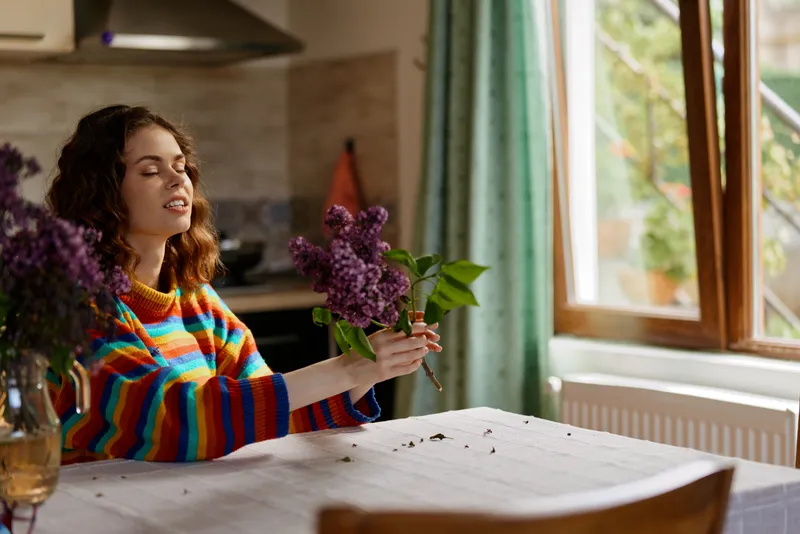 Cheerful woman indoors arranging a bouquet on a sunlit dining table, colorful striped sweater, relaxed pose, soft breeze through window, warm natural tones, cozy home mood