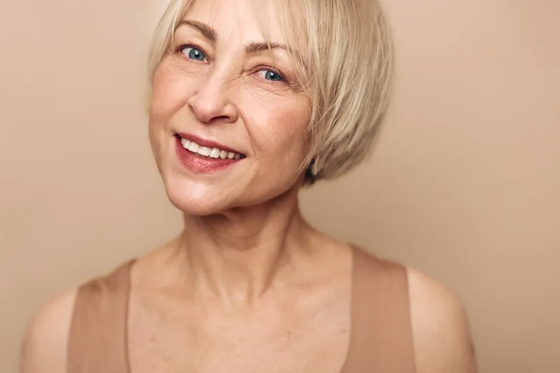 Closeup portrait of happy mature woman with short blonde hair smiling gently, natural skin texture visible, beige background. Concept of aging beauty, confidence, and wellness.