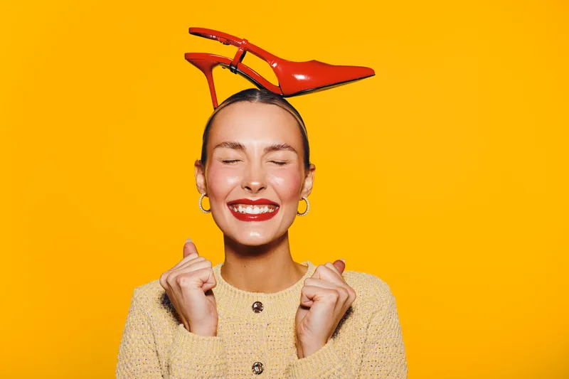 Happy woman with closed eyes smiles brightly wearing red high heel shoes on her head against yellow background. Concept of fun, fashion, joy, and playful mood in studio.