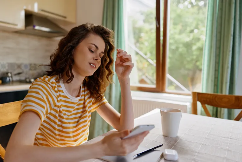 A woman sits at a bright kitchen table, absorbed in her smartphone, in a casual home moment. A calm, everyday scene suggesting relaxed work life and personal routine.