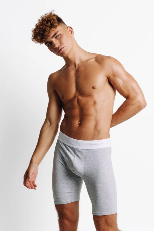 Young man with muscular body posing in gray shorts against a plain wall. Fit model with curly hair showcases toned abs and defined torso in clean, minimal studio setting.