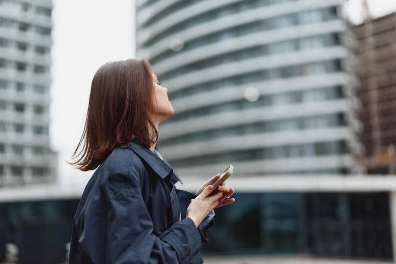 Young woman in a navy coat looks up while using a smartphone in an urban setting with modern buildings behind, creating a focused moment in city life