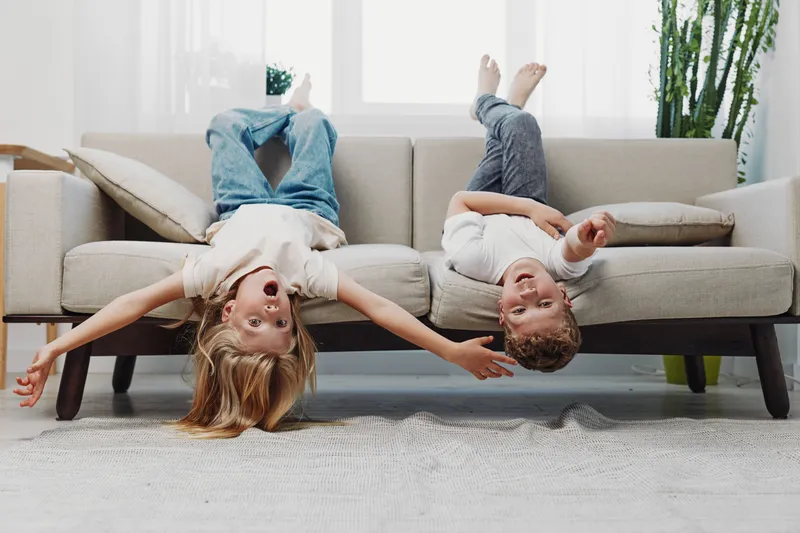 Two happy children lying upside down on a sofa with arms stretched out, laughing and playing in a bright living room with plants and cushions.