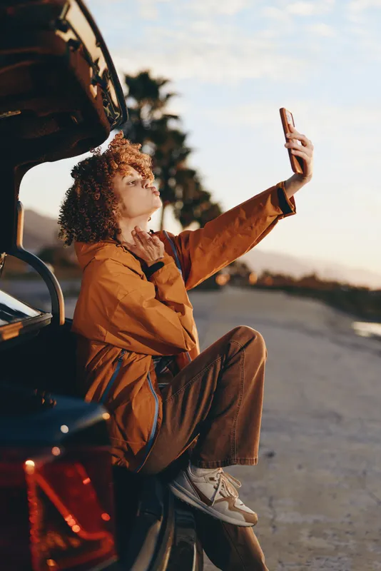 Woman sitting in car outdoors at sunset, holding a phone, relaxed lifestyle moment, curly haired in orange jacket, open door view, warm communication during a selfie