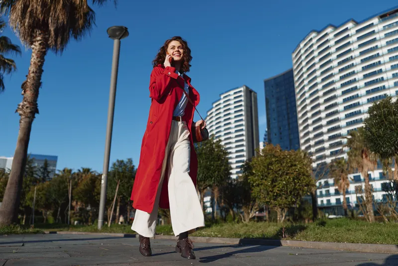 Young woman in a red coat walks confidently along an urban street with modern buildings and palm trees in the background on a sunny day.