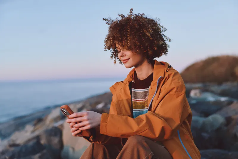 Woman lifestyle outdoors by seaside, holding a smartphone, curly hair in wind, wearing an orange jacket, calm smile at sunset, rocky shoreline, natural colors, soft light