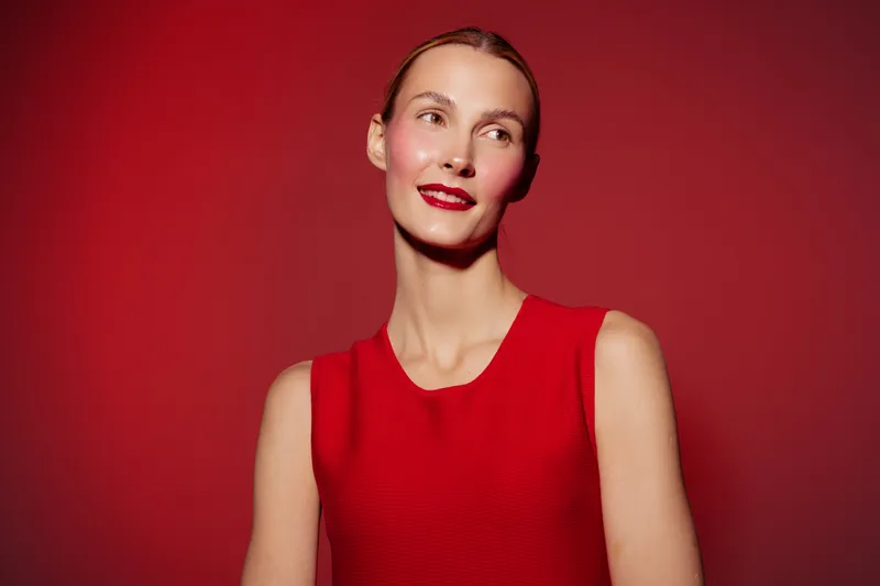 Young woman in red sleeveless dress poses against red background, smiling softly. Concept of fashion, beauty, elegance, and confident female model with natural makeup and red lipstick.