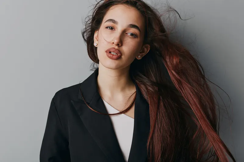 Young woman with long brown hair and natural makeup wearing black blazer and white top posing against gray background for fashion portrait and beauty concept.
