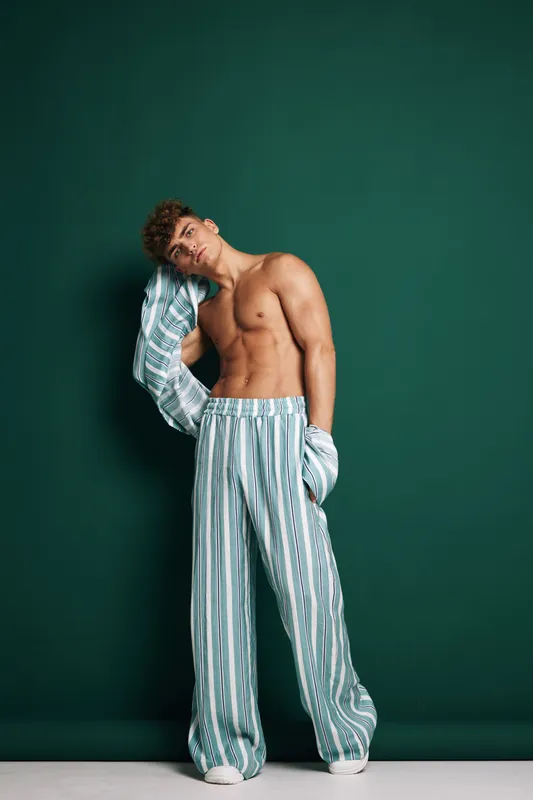 Young man, shirtless, posing with relaxed jacket over shoulder standing against green background. Trendy lifestyle, casual fashion, model, confident, modern style, barefoot in striped loose pants.