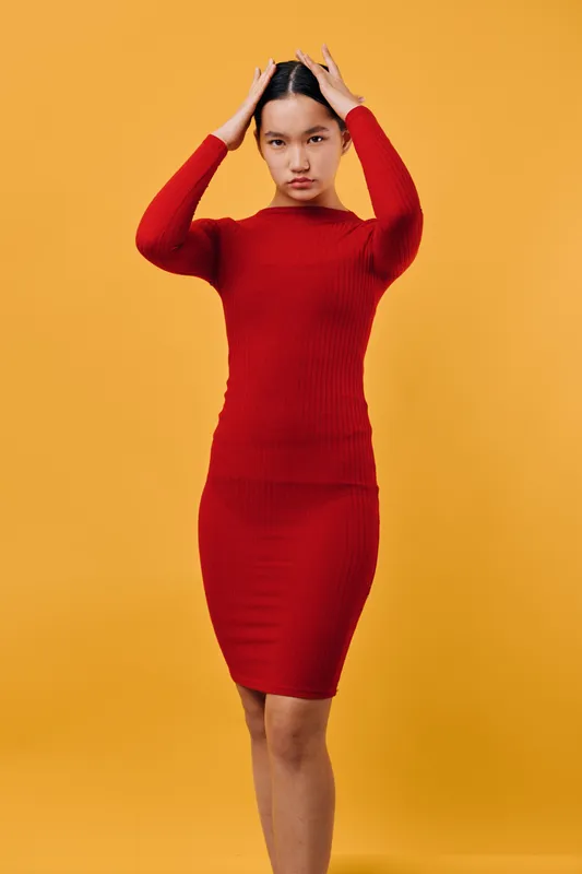 Young woman in red knitted dress posing with hands on head against orange background. Fashion model with serious expression standing in casual posture for portrait photography.